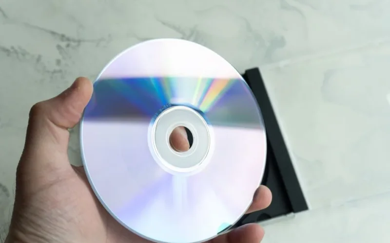 Close-up of a person holding a reflective CD disc with a transparent case on a marble background.
