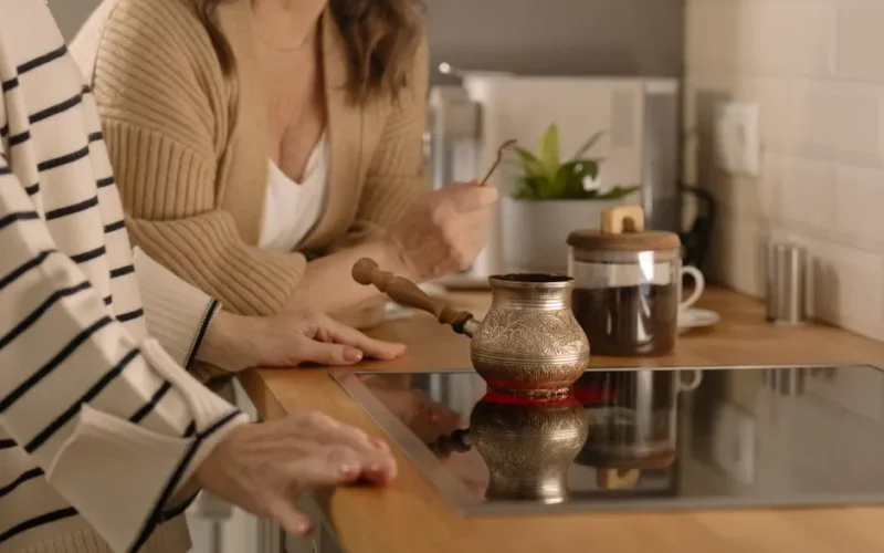 Two women making coffee on an induction stove, featuring a traditional coffee pot and jar on a wooden kitchen counter.