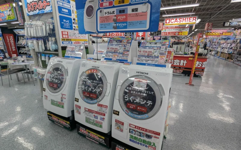 Several Hitachi drum washing machines lined up in a Japanese electronics shop, each bearing promotional materials for easy maintenance.