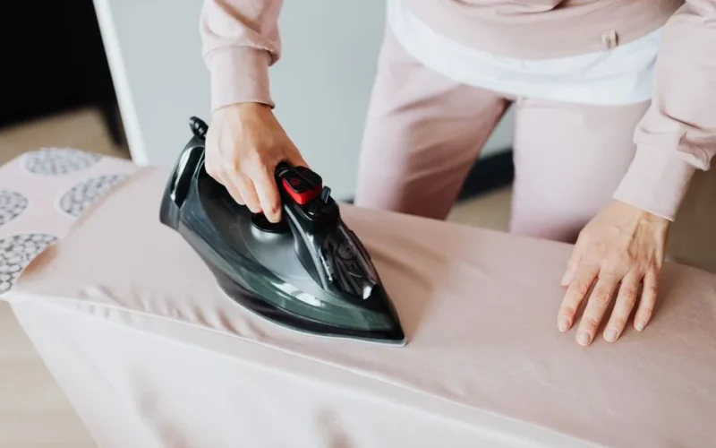 Adult woman ironing clothing on board using a black iron in an indoor setting.