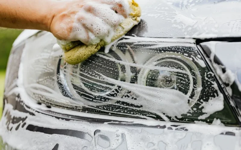 Hand scrubbing a car headlight with sponge and soap suds, detailing process.