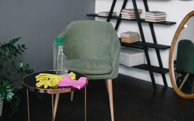 Yellow latex gloves placed near spray detergent bottle and rag on round table against comfortable chair in living room in daytime