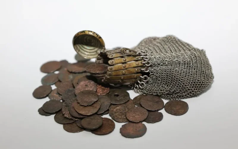 Antique metal chain pouch spilling ancient rustic coins on a white surface.