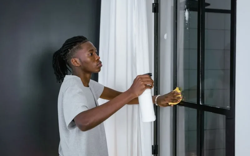 A man cleaning a glass door indoors using a spray bottle and cloth.