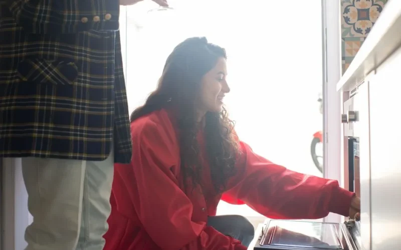 Woman in a red shirt operating an oven indoors, smiling in a bright kitchen environment.