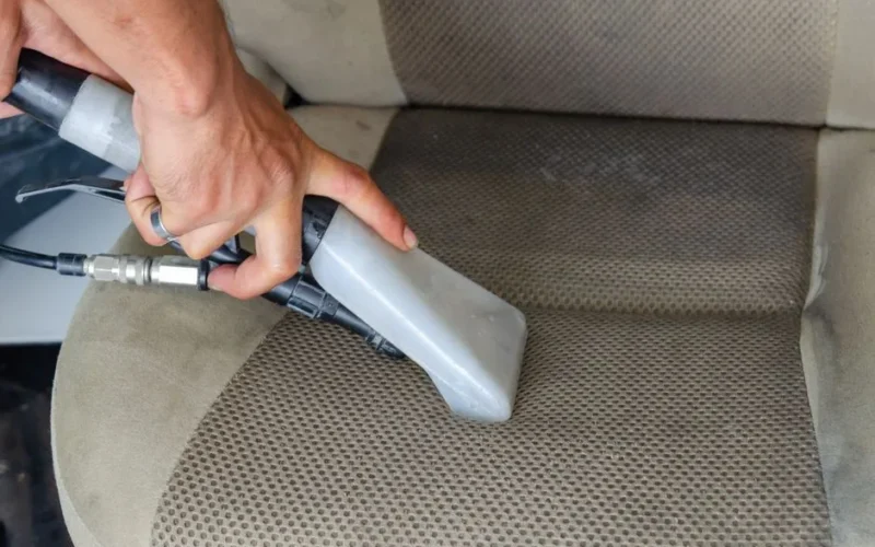 Hands using a vacuum nozzle to deeply clean a fabric car seat, removing dirt and stains.