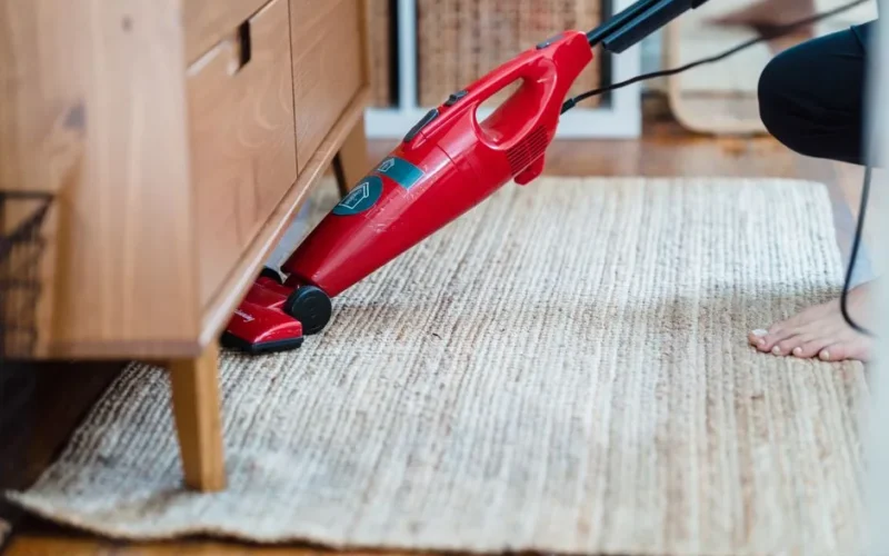 Side view of a person using a red vacuum under furniture on a rug for effective cleaning.