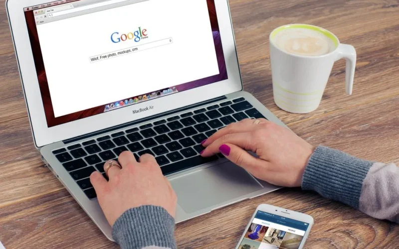 Hands typing on MacBook Air with Google search open, coffee nearby.