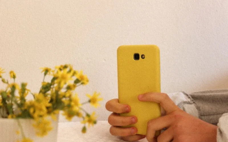 Hands holding a smartphone with a yellow case next to a vase of flowers indoors.