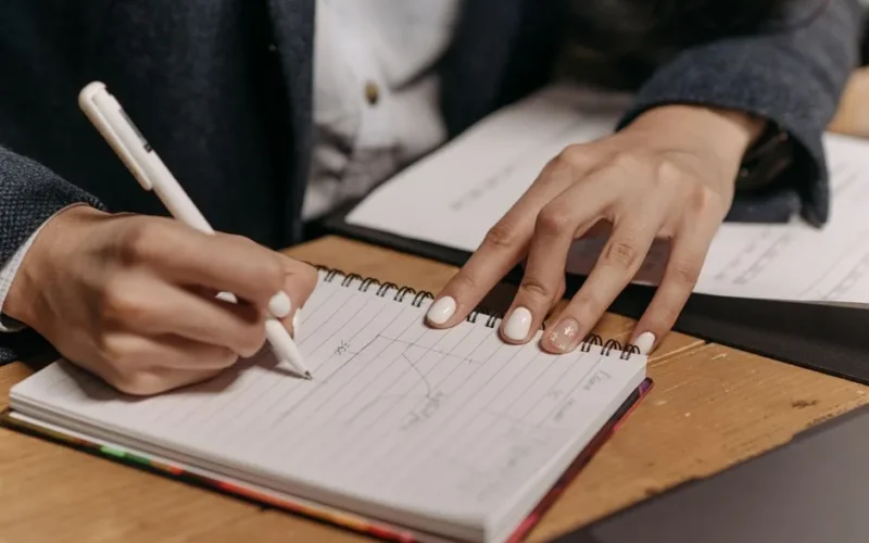 Close-up of a person writing notes in a spiral notebook with a pen on a wooden desk.