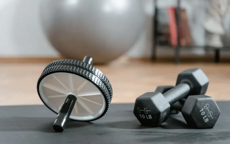 Close-up of ab wheel and dumbbells on a mat in a home gym setting.