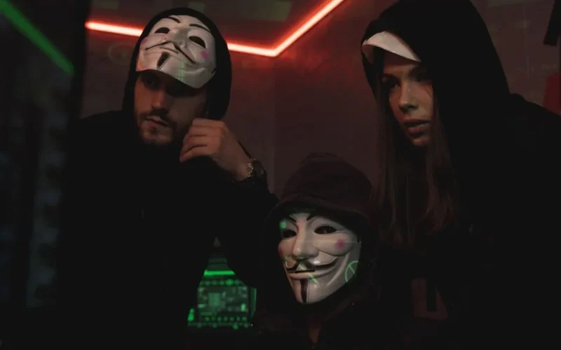 A group of people wearing Guy Fawkes masks in a dark tech-themed room, suggesting anonymity and hacking activities.