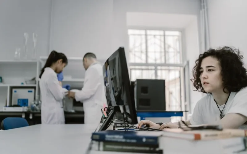 Focused woman working on a computer in a busy laboratory setting, showcasing teamwork and scientific research.