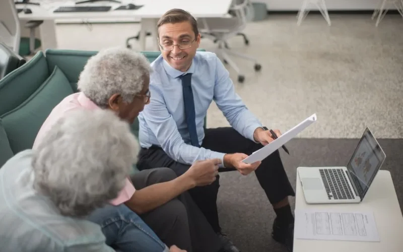 Consultant discussing financial plans with senior clients in a modern office setting, using documents and a laptop.
