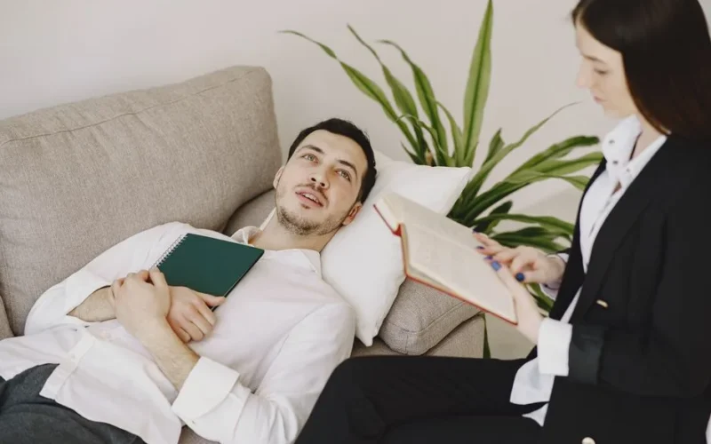Thoughtful well dressed man with journal lying on sofa in office of modern female therapist and telling about problems