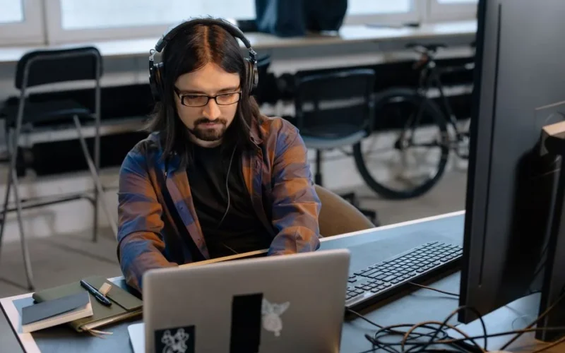 Bearded male developer in headphones coding on a laptop in a modern office setting.