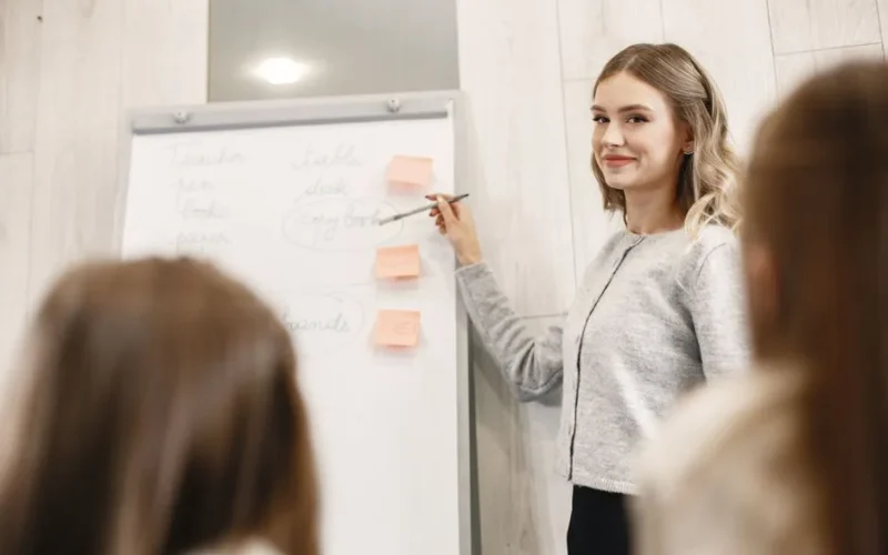 Female teacher smiling as she explains notes on a flipchart to a class.
