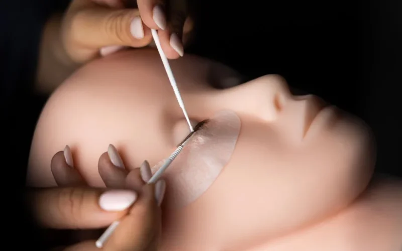 Close-up of a beautician practicing eyelash extensions on a mannequin head with precision and focus.