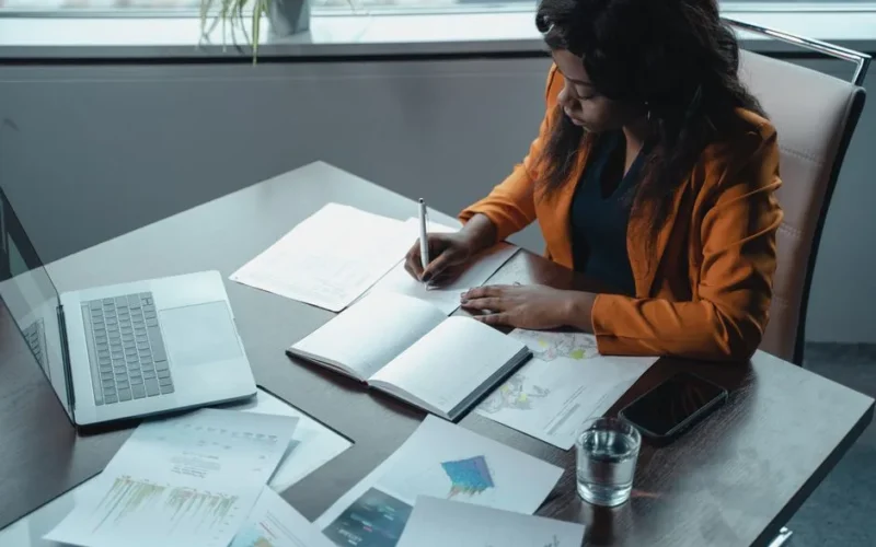 Businesswoman in office writing notes with documents and laptop, focused on analysis and productivity.