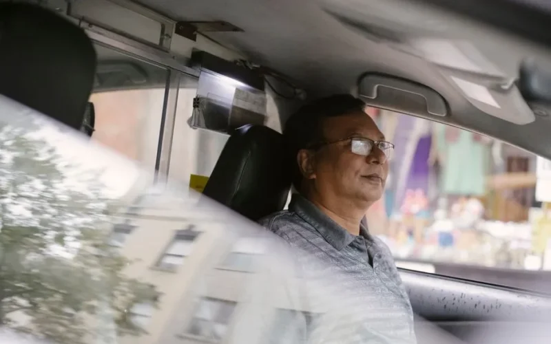Side view of Asian middle aged male in eyeglasses sitting on driver seat in automobile with reflection of building on half opened window
