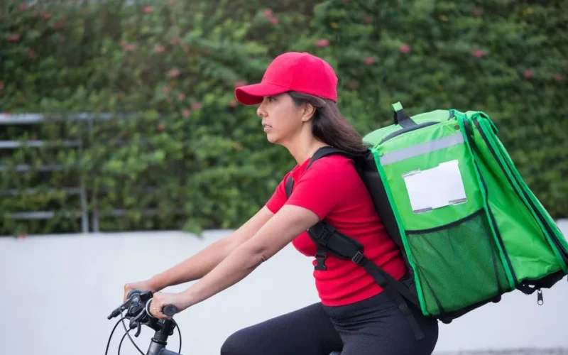 A female courier wearing a red cap rides a bicycle with a large green delivery bag outdoors.