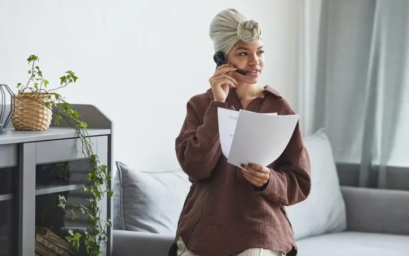 African American female call agent with headset and documents sitting on couch during conversation in light room with cupboard and plant