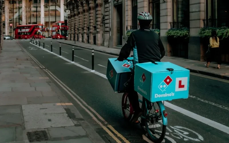 Delivery person on a bicycle in urban city with iconic red double-decker buses and historical buildings.