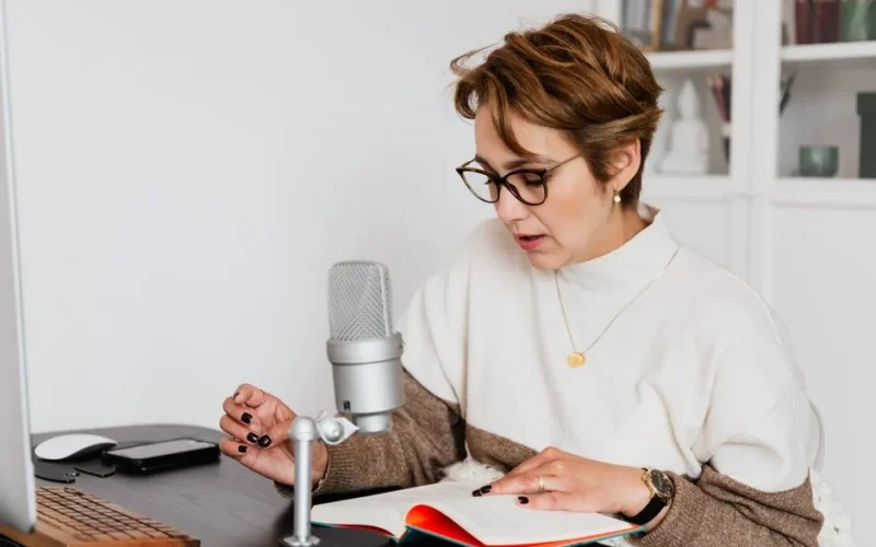 Female narrator in glasses reading out loud from book while sitting at desk with microphone and recording audiobook in cozy study