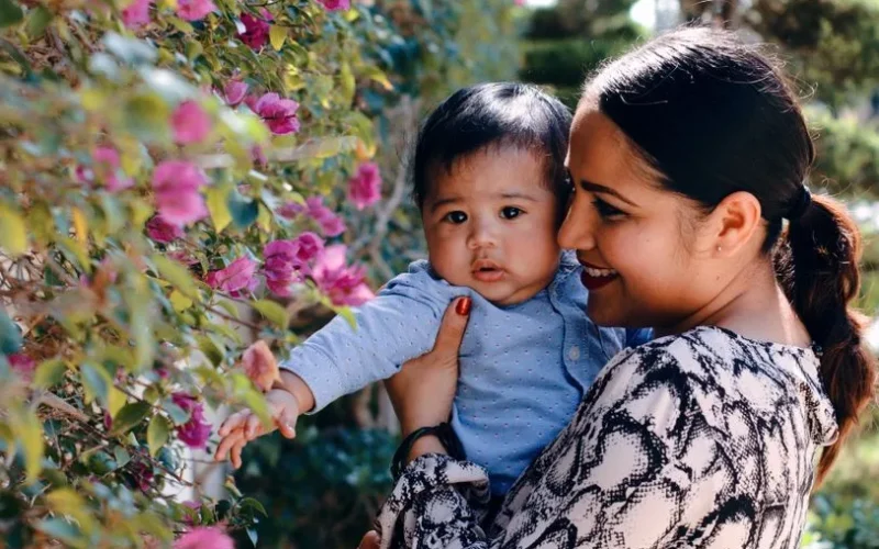 Cheerful young ethnic female hugging cute baby boy and smiling while spending time together in garden near flowers