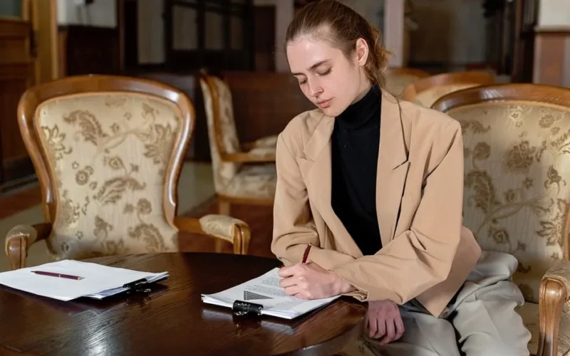 Young woman in a brown blazer signing documents at a table in a stylish office.