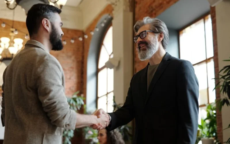 Two businessmen smiling during a handshake in a modern office setting, symbolizing success and partnership.
