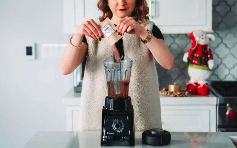 A woman pours a sachet into a blender in a festive kitchen setting.