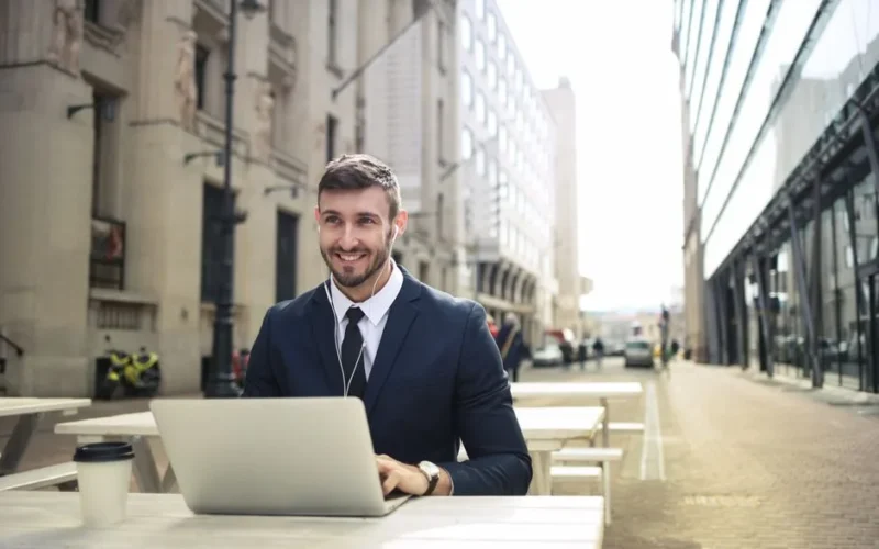 Confident businessman in formal wear using a laptop outdoors in a city setting on a sunny day.