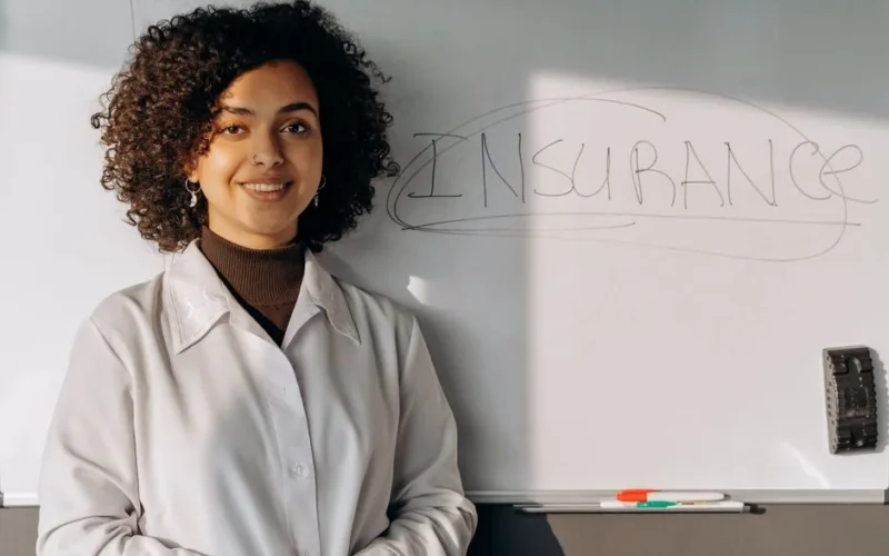 African American woman smiling in office setting with a whiteboard displaying 'Insurance'.
