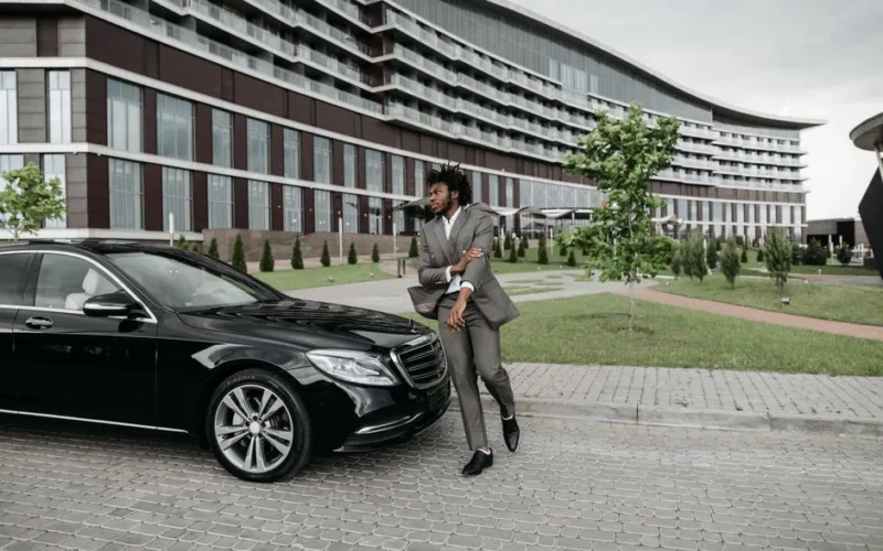 Confident man in suit poses beside luxury car with modern building backdrop outdoors.