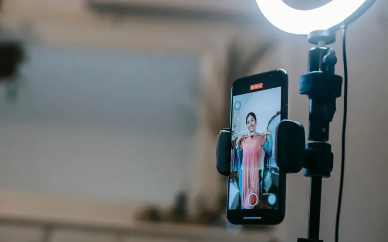 Anonymous female blogger standing in room and shooting vlog on smartphone with ring lamp while demonstrating pink dress in hands