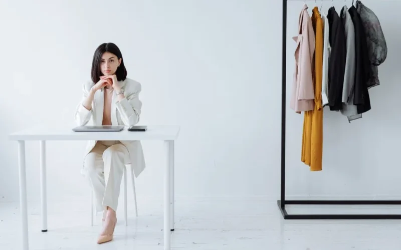 Modern businesswoman sitting at desk with clothing rack, embodying fashion and professionalism.