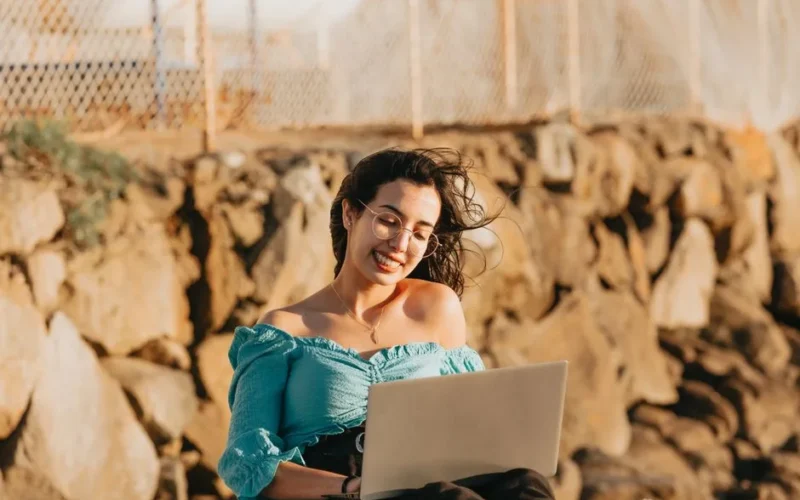 Woman enjoying remote work with laptop outdoors, smiling on a windy day.
