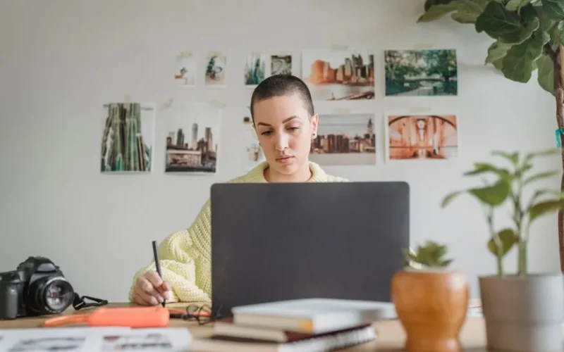Professional female designer looking at laptop and using graphic tablet while sitting at table in light workspace