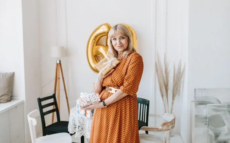 Elderly woman in orange dress holding gifts and smiling, celebrating birthday indoors.