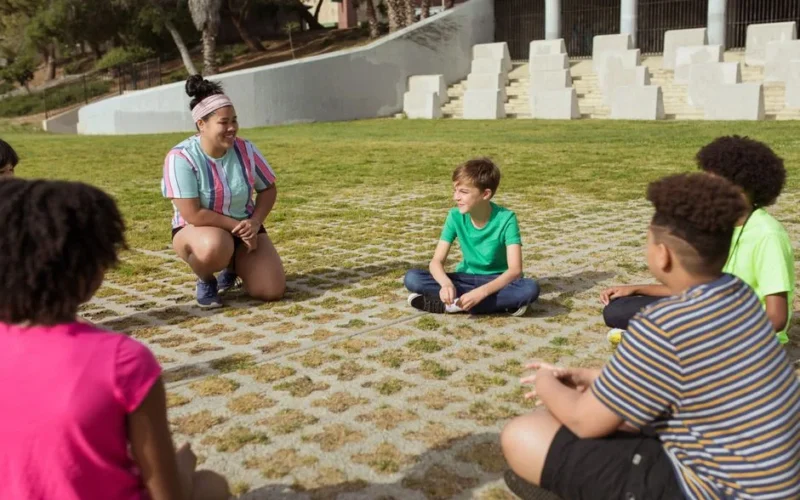 A diverse group of kids sitting in a circle outdoors, engaged in a fun summer camp activity.