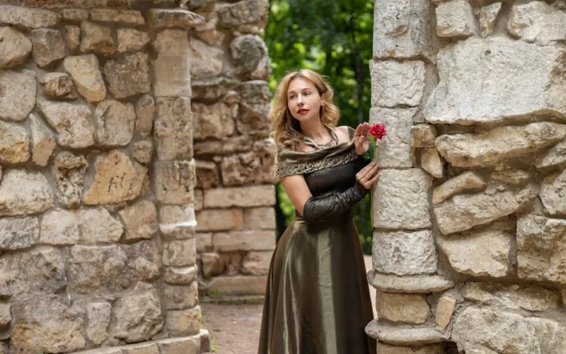 Woman in medieval dress with a rose amidst stone ruins, exuding timeless beauty.