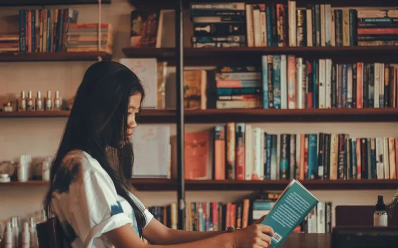 A young woman reads a book in a comfortable library with shelves full of books, offering a serene setting for relaxation.