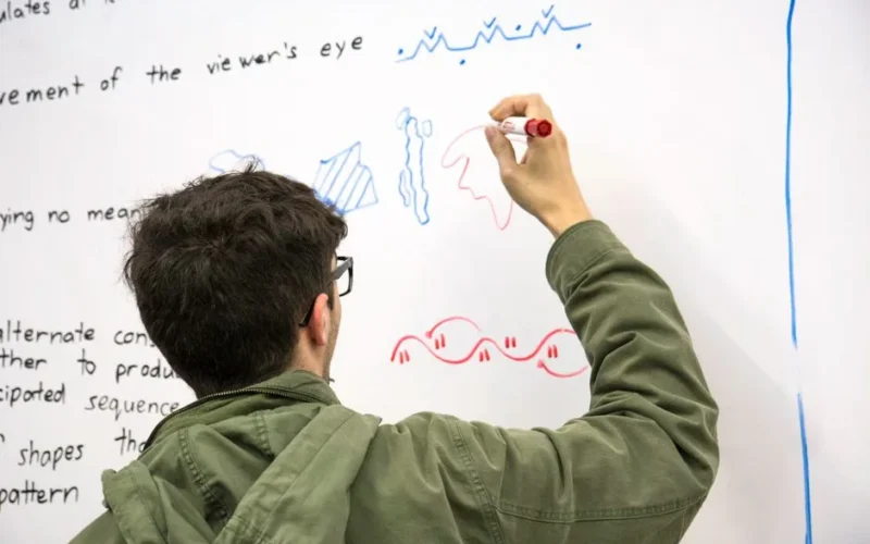 A student in a green jacket draws colorful diagrams on a whiteboard, illustrating educational concepts.
