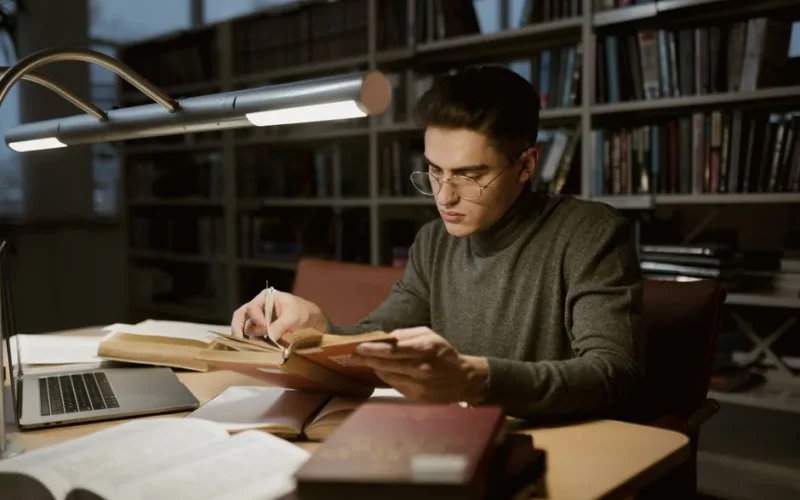 A young man in a sweater reads books in a library, surrounded by books and a laptop.