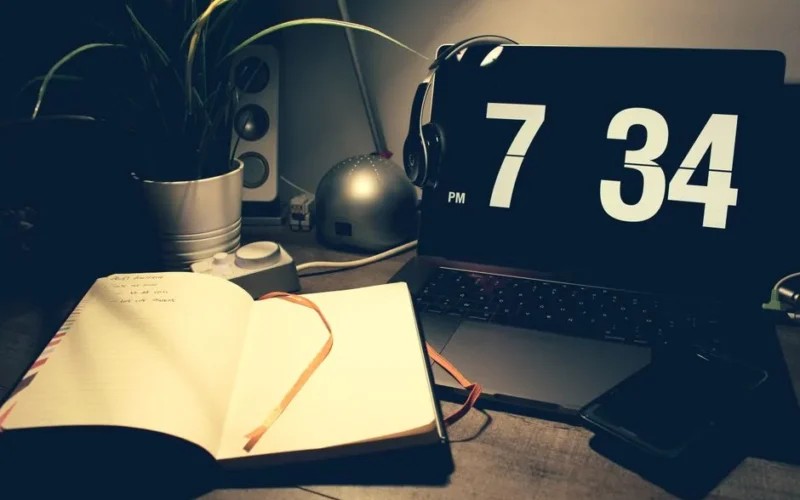A cozy workspace with a laptop displaying time, open notebook, and headphones under warm lighting.