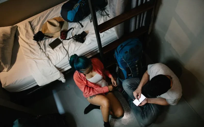 A young couple using a smartphone in a hostel room with backpacks, ready for travel.