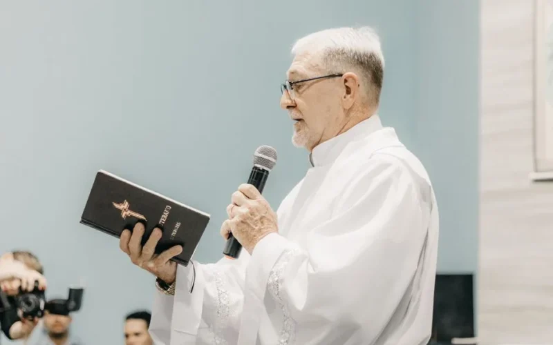 Priest in white robe holding Bible and microphone during church ceremony.