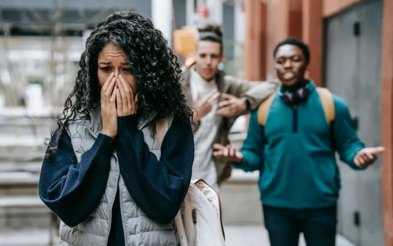 Diverse young male friends scoffing at ethnic female groupmate crying and covering mouth with hands on street