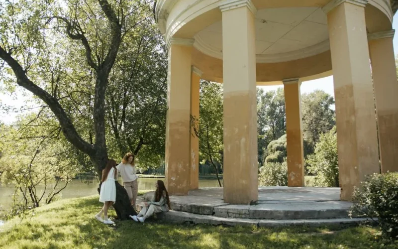 Three women enjoying a serene day by a gazebo in a lush, sunlit public park.
